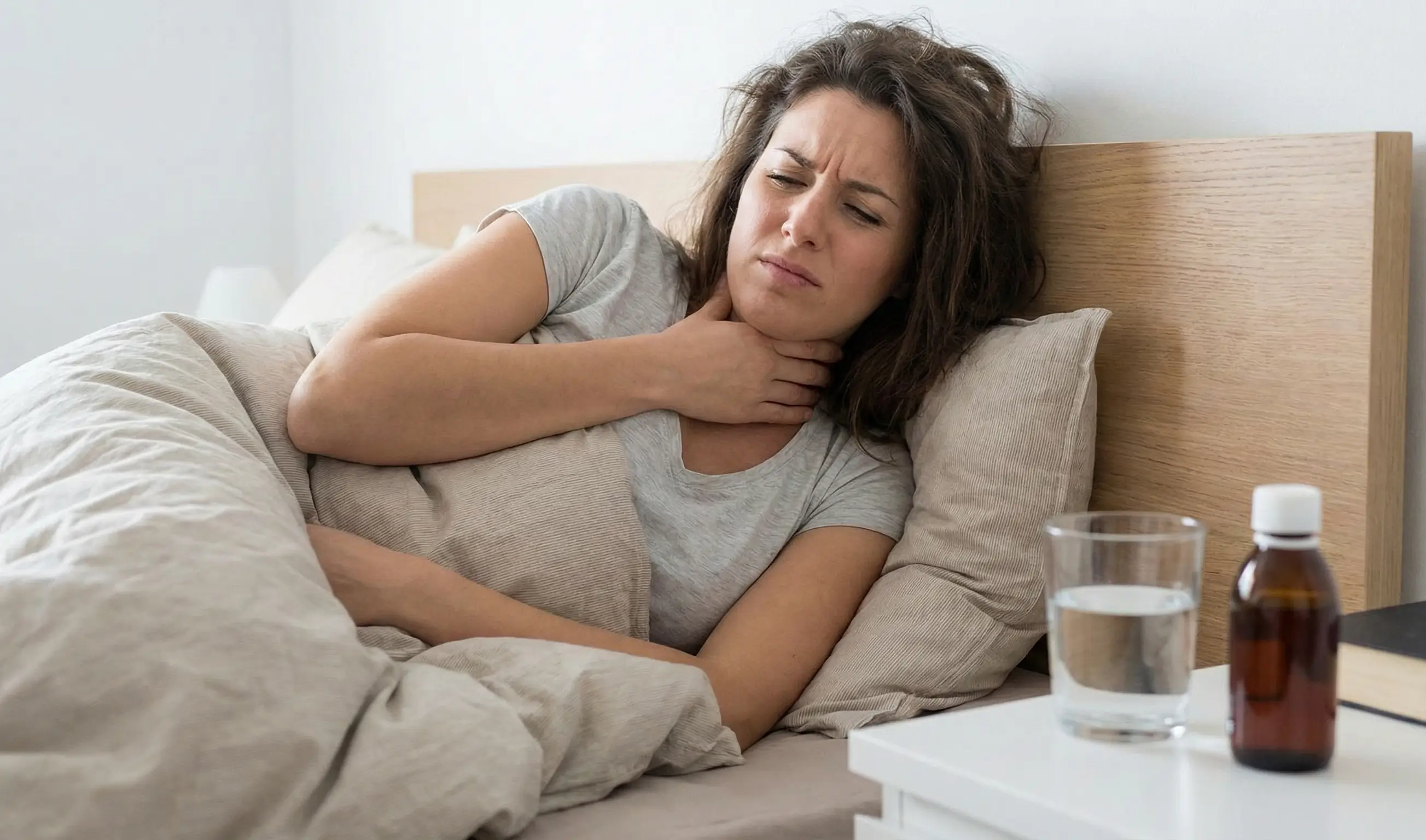 Femme malade au lit avec mal de gorge, main sur la gorge, verre d'eau et médicaments sur la table de nuit